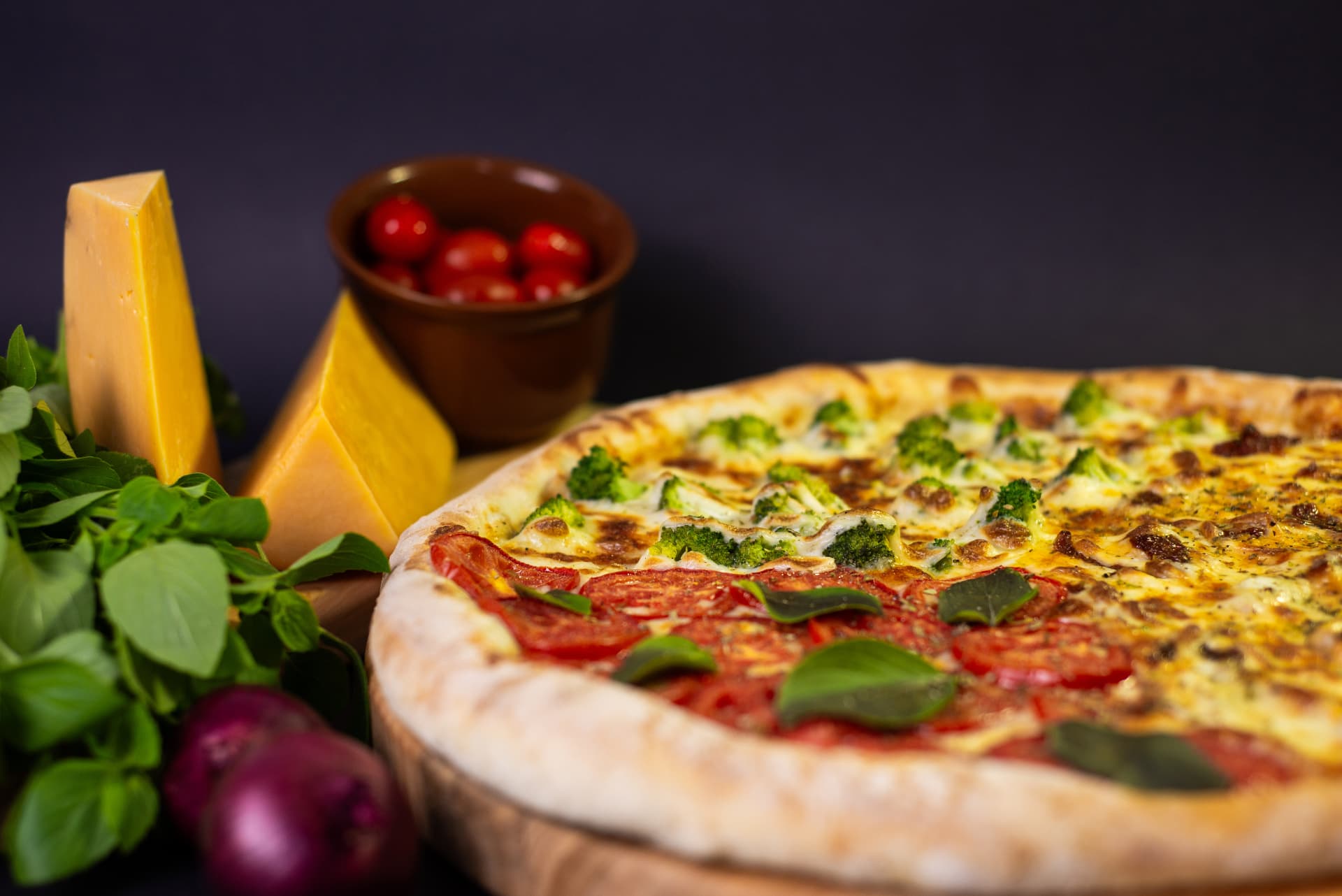 Pizza com queijo, tomate e brócolis sobre mesa de madeira, ao lado de queijos, ervas e tomates