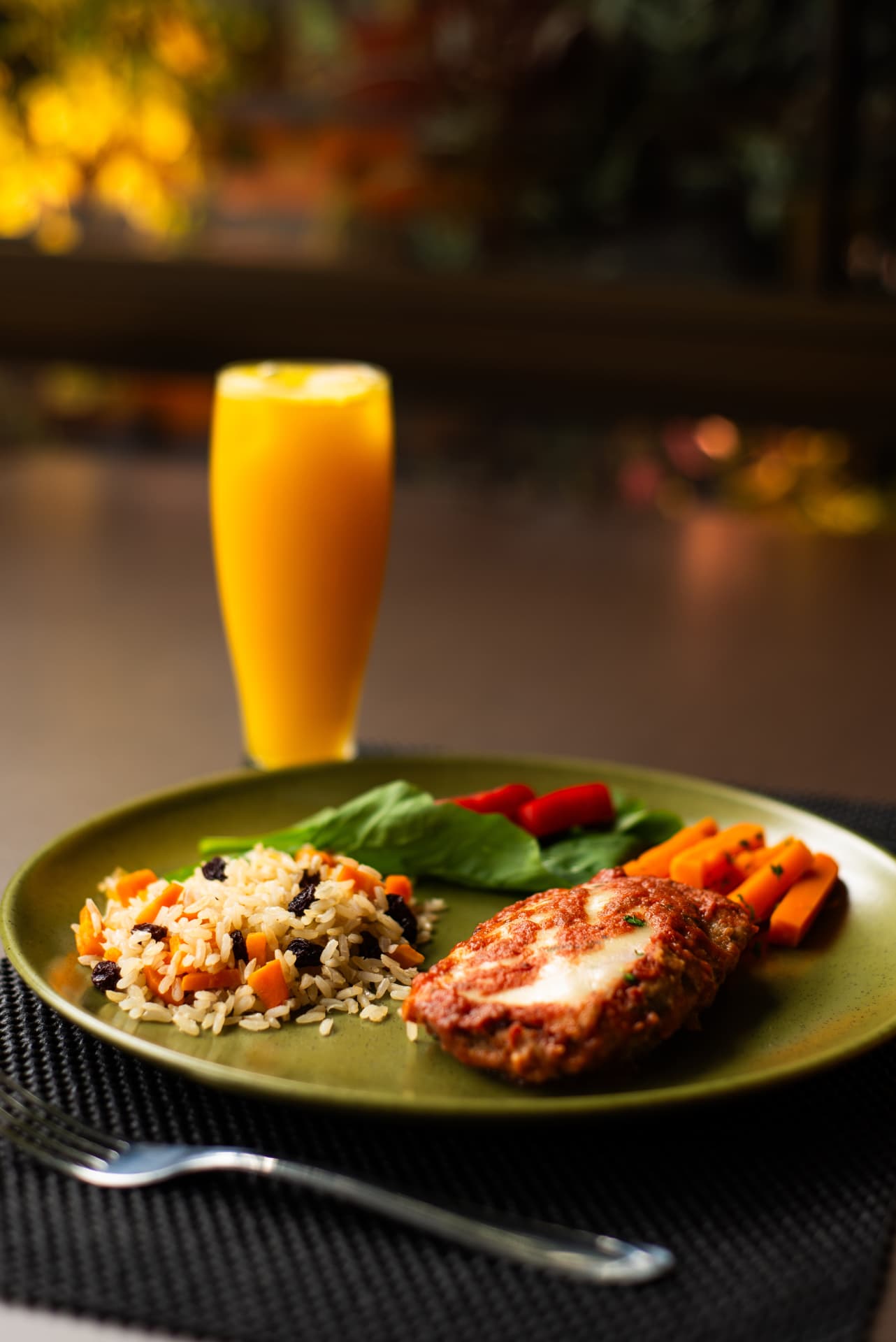 Prato verde com filé empanado, arroz, folhas e cenouras, acompanhado de um copo de suco de laranja